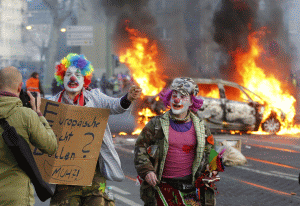 Protestatari îmbrăcaţi în clovni trec pe lângă o maşină de poliţie în flăcări (Foto Mediafax)