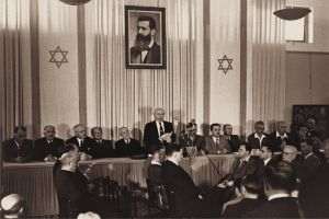 David Ben-Gurion (the first Prime Minister of Israel), publicly pronouncing the Declaration of the State of Israel, on May 14, 1948 (Tel Aviv, Israel), under a large portrait of Theodor Herzl, the founder of modern political Zionism, in the old building of the Tel Aviv Museum of Art. (Photo source: Israel Ministry of Foreign Affairs, Author: Rudi Weissenstein)